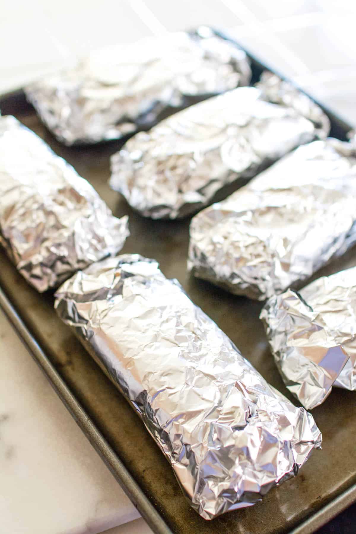 A cookie sheet of sandwiches wrapped in aluminum foil.