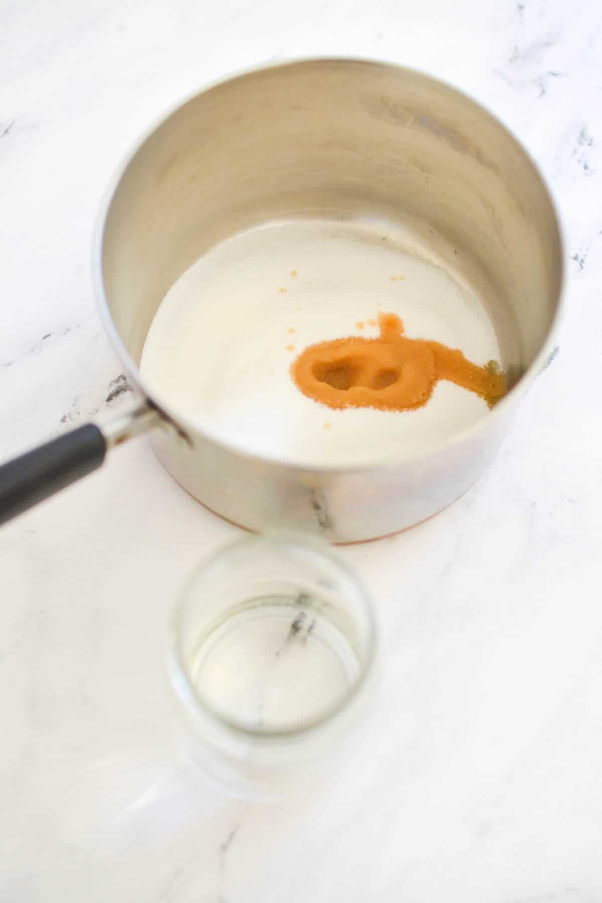 A saucepan with white sugar and vanilla extract next to a jar of water.