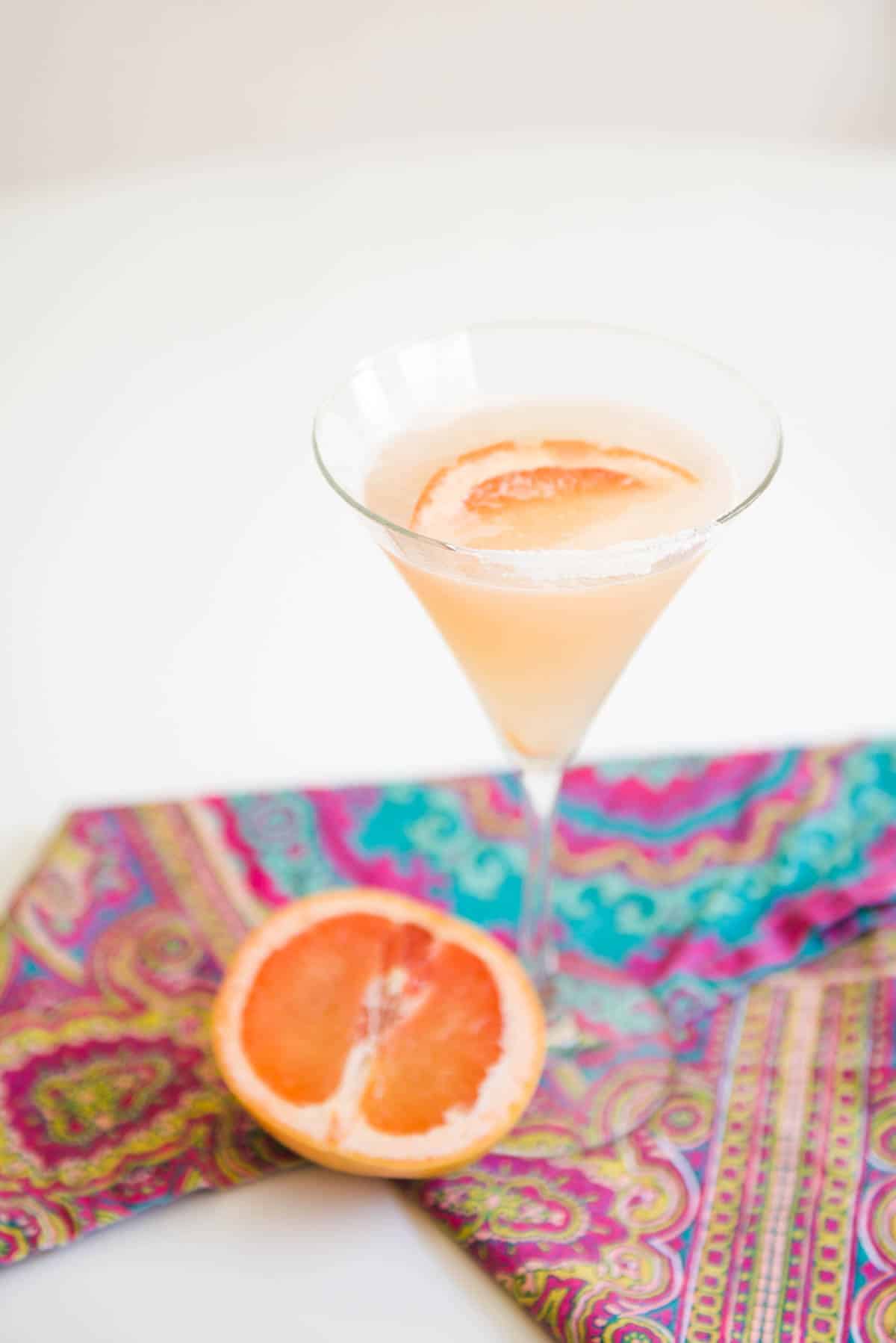 Martini glass on a table with a grapefruit martini in it with a fresh grapefruit on the table next to it.