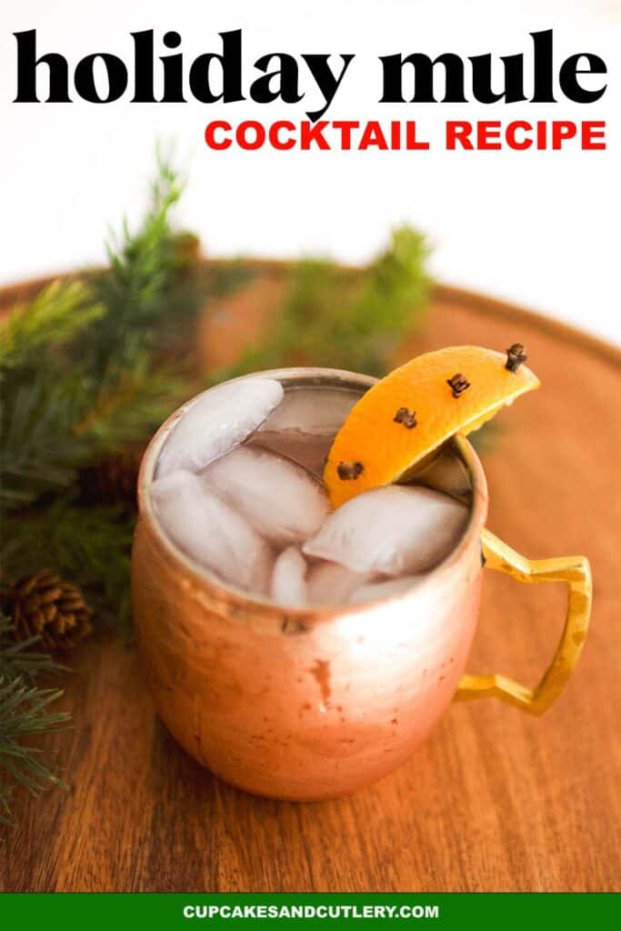 Image of a holiday Moscow Mule garnished with a clove studded orange slice on a wood cutting board. Text reads, Holiday mule cocktail recipe.