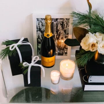 A bottle of champagne next to candles and some Christmas gifts on a table.