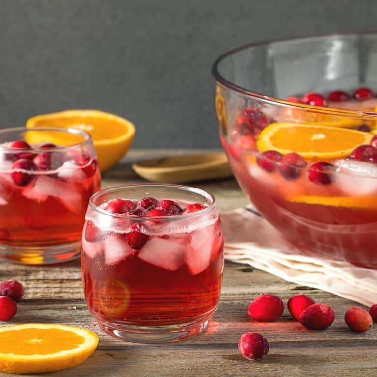 A glass punch bowl with a red punch in it next to a glass of punch.