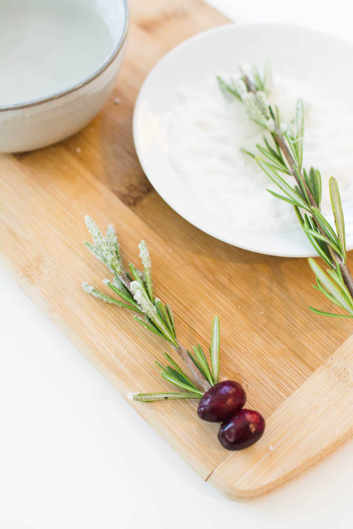 A sprig of sugared rosemary on a tray next to a plate of sugar with a sprig of rosemary.