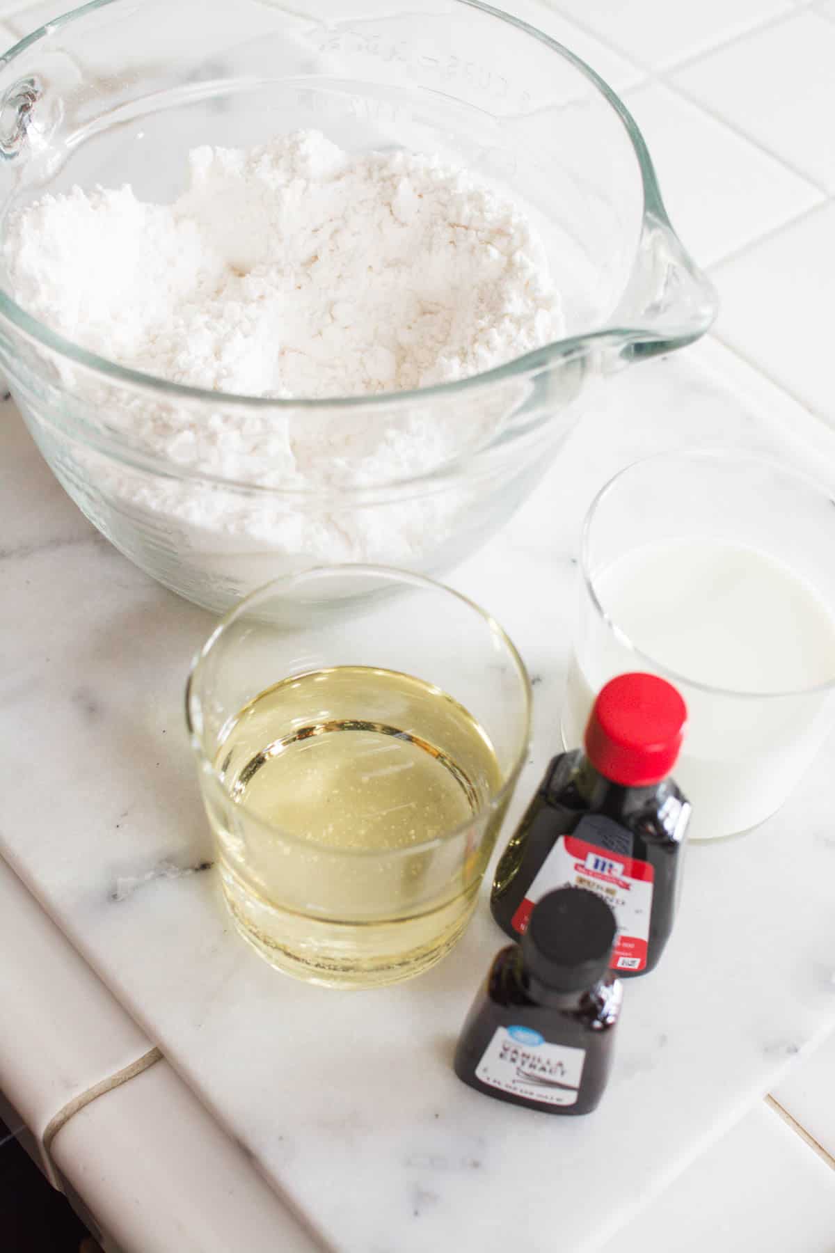 Dry cake mix in a glass bowl next to vanilla, oil and almond oil on a counter.