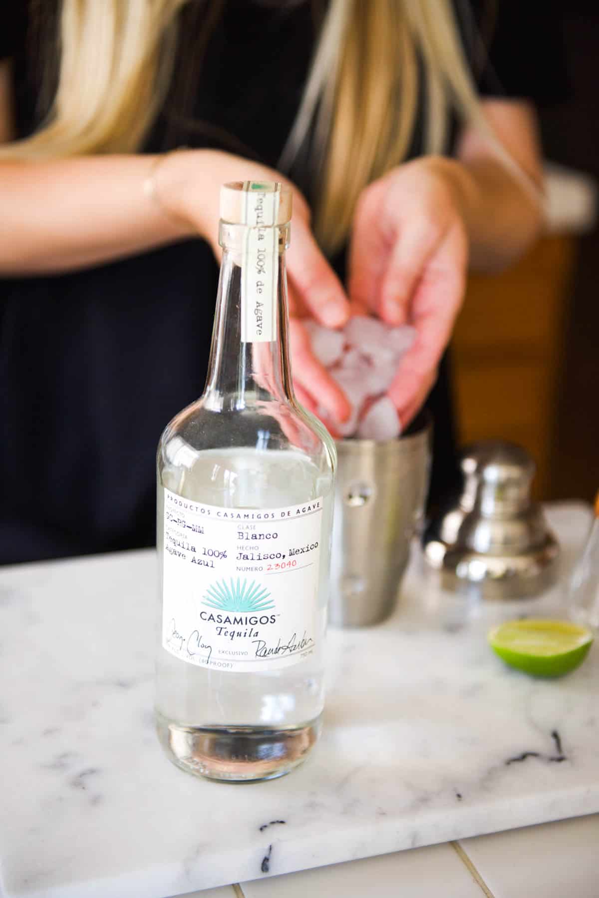 A bottle of tequila on a counter with a woman making a cocktail behind.