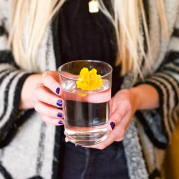 A glass of water with a yellow flower in it in a glass held by a woman.