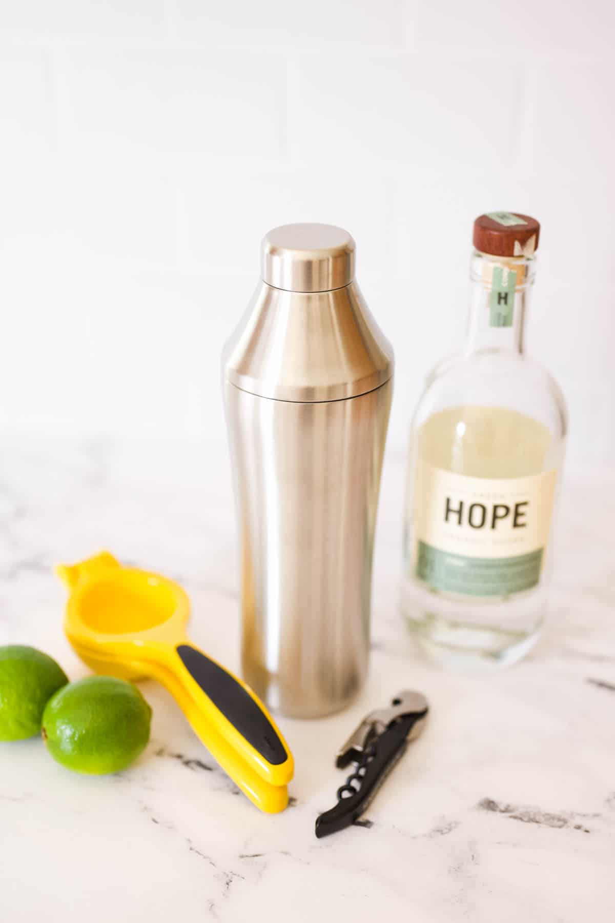 A bottle of vodka, cocktail shaker, limes and a citrus squeezer on a table.