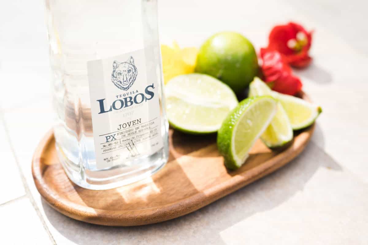 Close up of a bottle of Lobos tequila on a wooden tray next to limes and flowers.