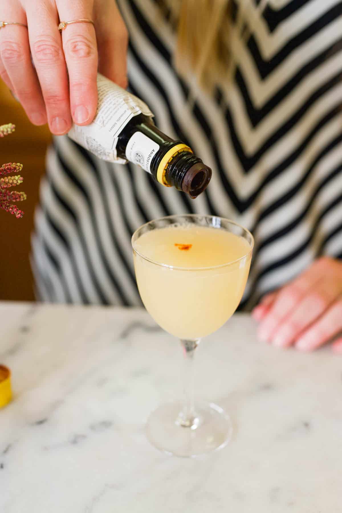 Woman adding drops of cocktail bitters to the top of a lemon cocktail.