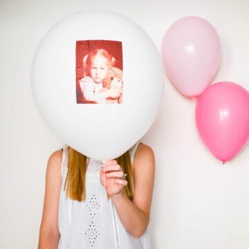 A woman holding a white balloon in front of her face with a photo of a little girl pasted on it.