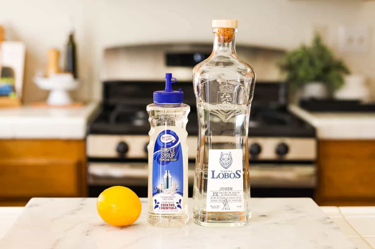 Bottle of tequila, fresh whole lemon and a bottle of simple syrup on a counter.