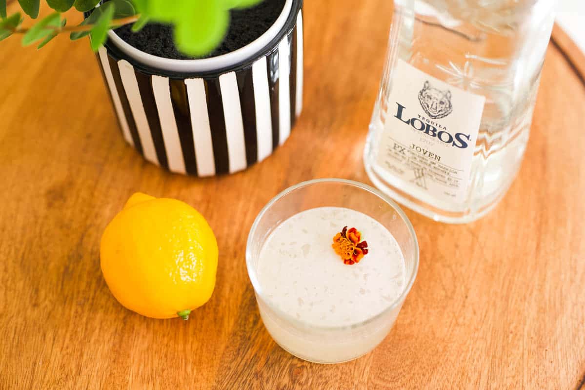 A cocktail on a tray with a lemon a bottle of tequila and a black and white striped planter.
