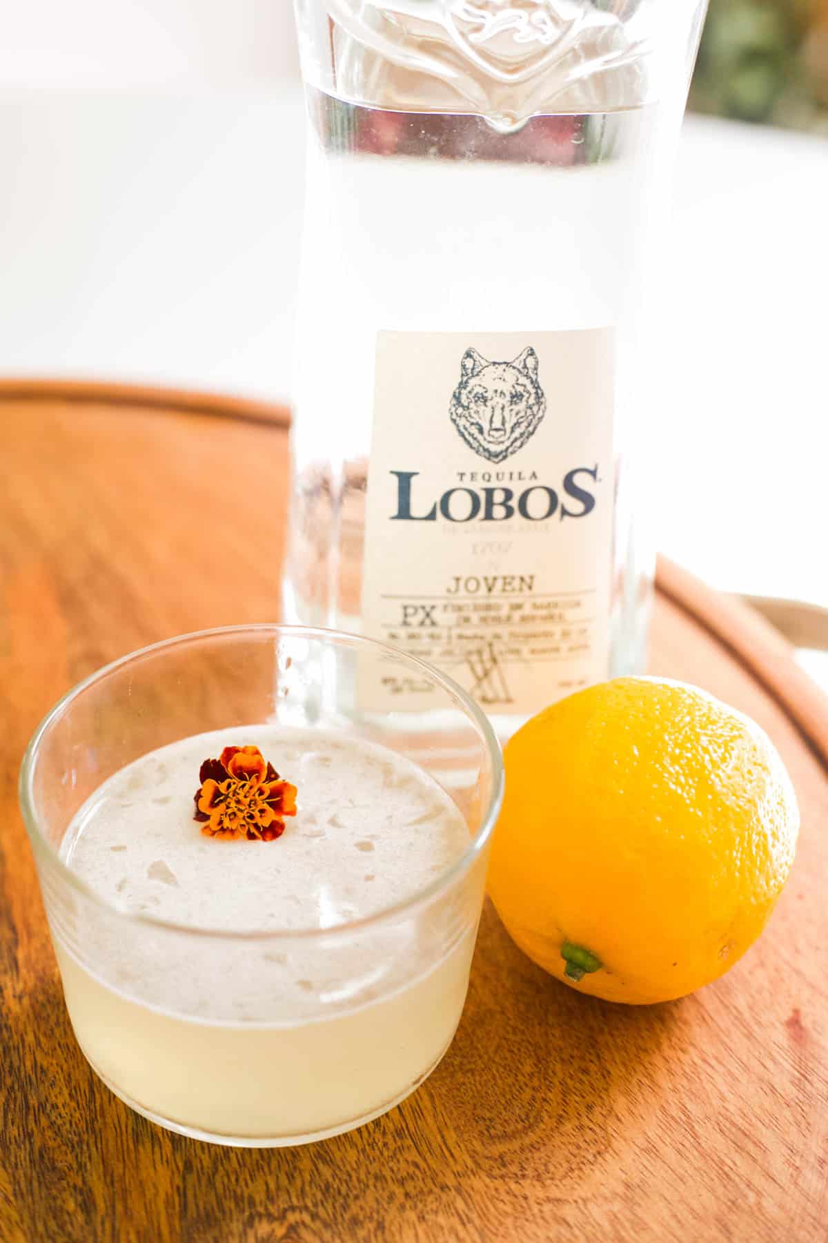 A glass with a tequila sour recipe in it next to a fresh lemon and bottle of tequila.