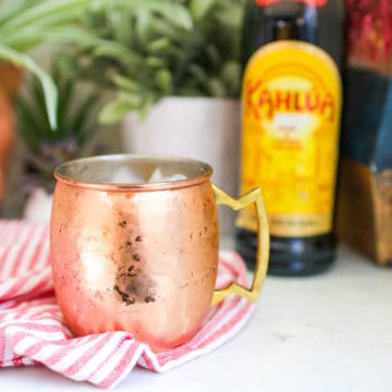 A White Russian Mule in a copper mug on a table in front of a bottle of coffee liqueur.