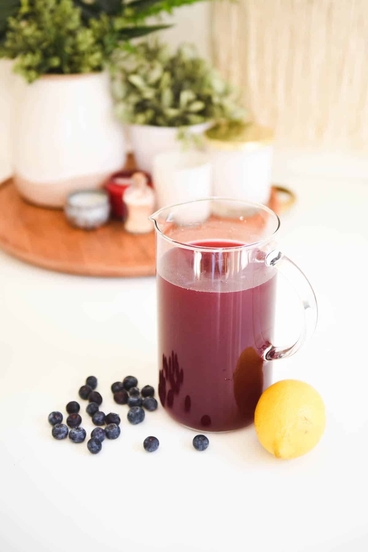 Fresh blueberries and a whole lemon on a table next to a pitcher of Blueberry Lemonade.