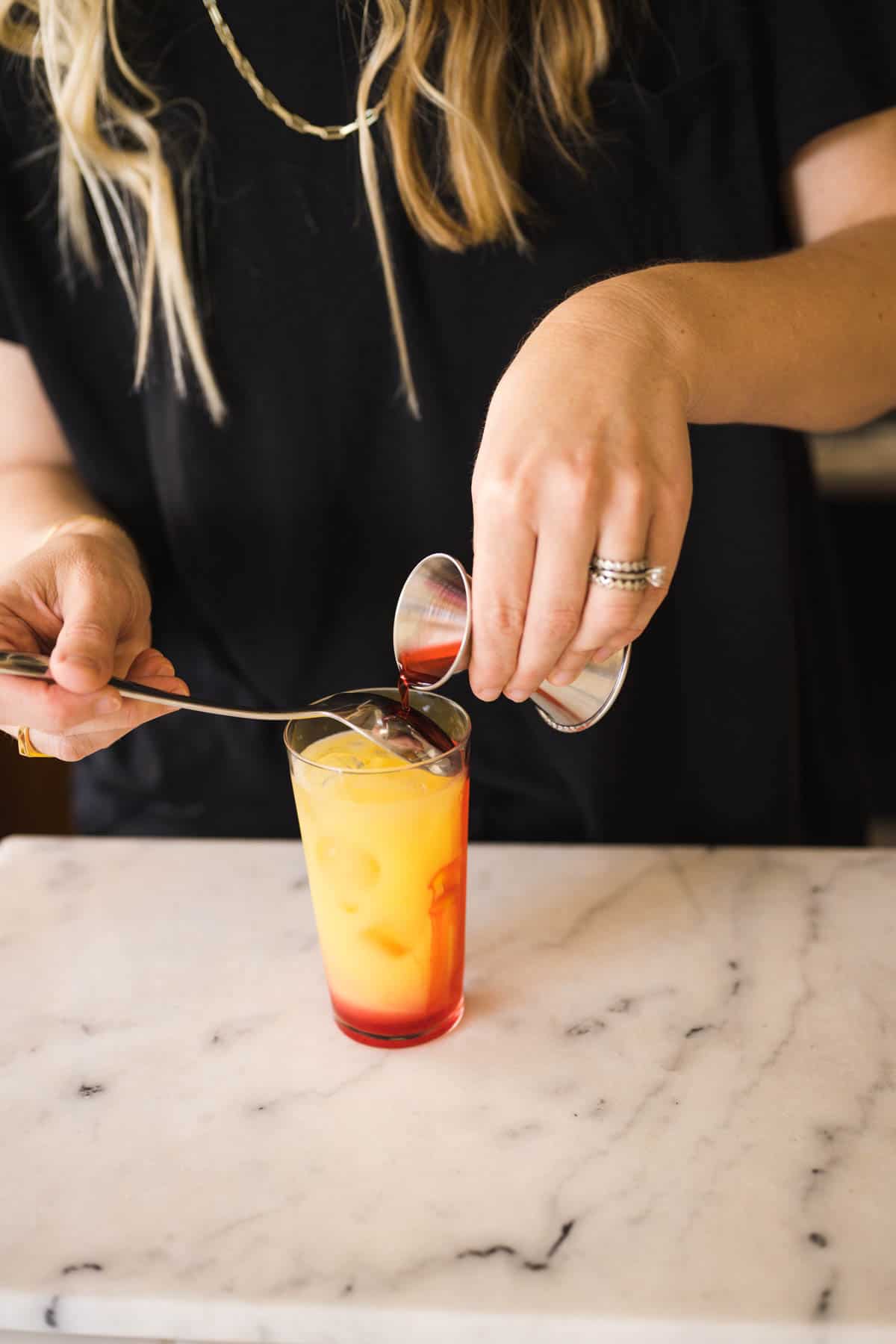 Woman pouring grenadine over the back of a spoon into a glass to make a Tequila Sunrise.
