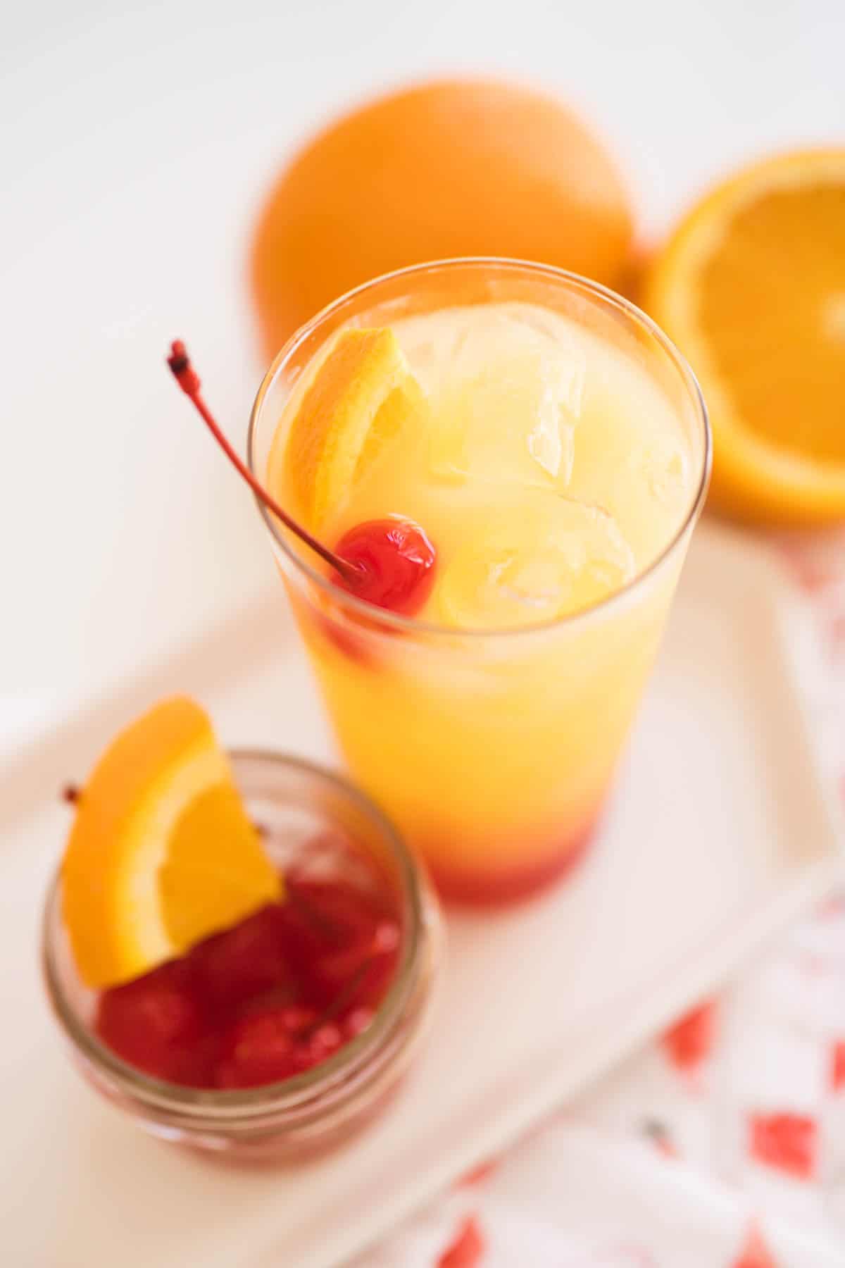 A finished Tequila Sunrise in a tall glass garnished with a piece of orange and a cherry.