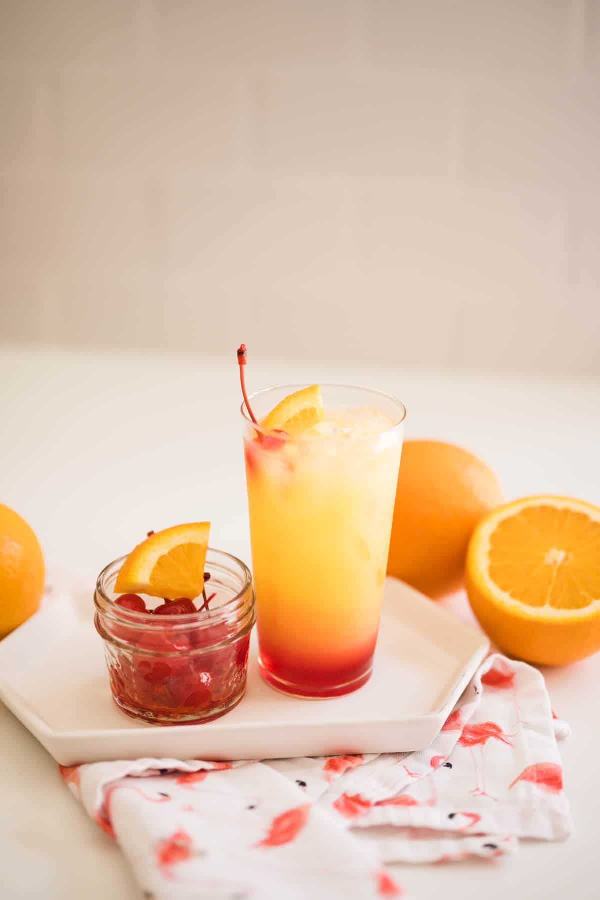A Tequila Sunrise cocktail on a table next to some oranges and a small bowl of cherries.