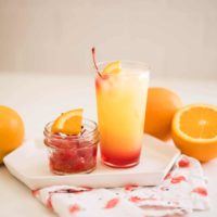 Close up of a Tequila Sunrise cocktail on a table next to a bowl of cherries and oranges.