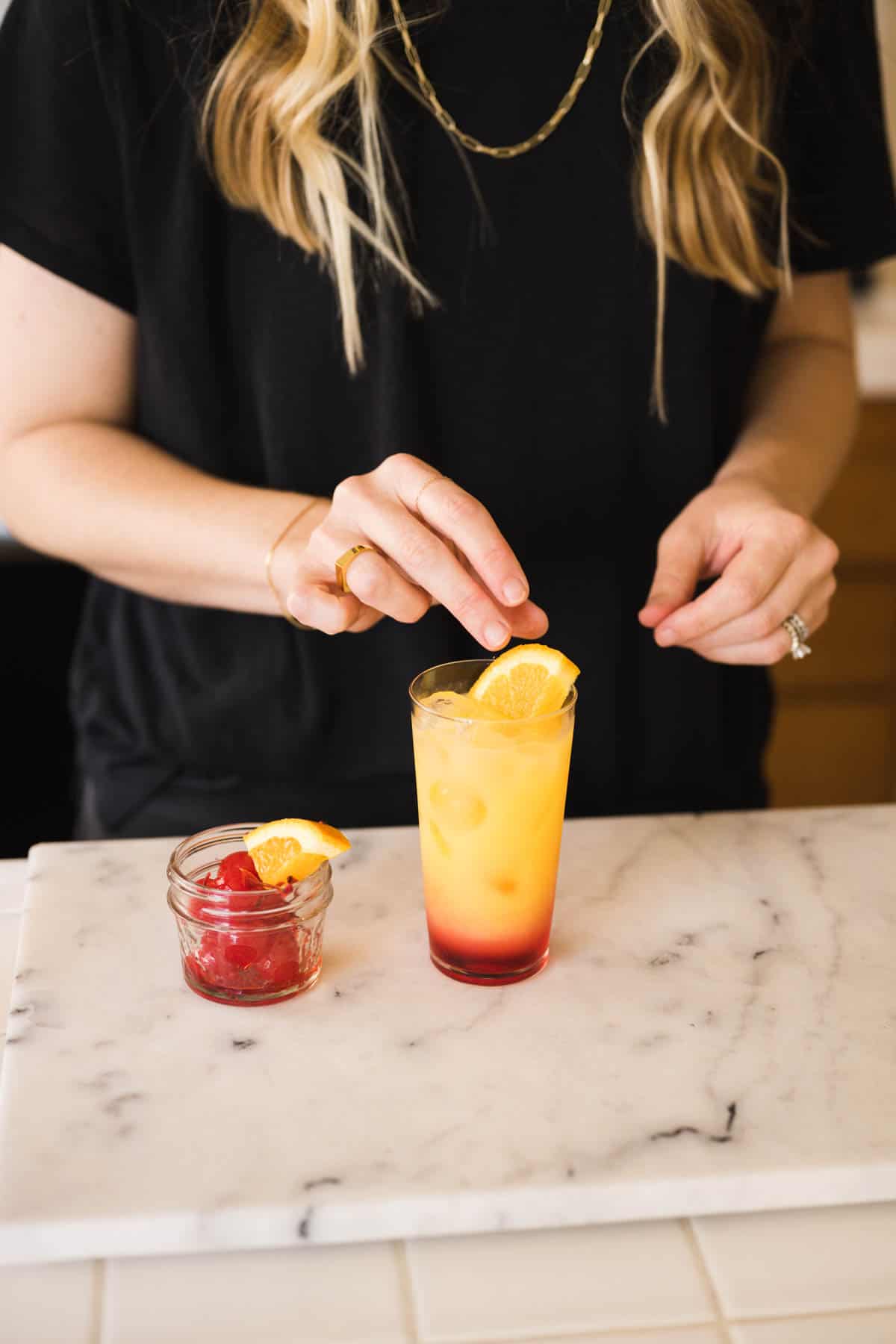 Woman adding a cherry and piece of orange to a finished Tequila Sunrise recipe.