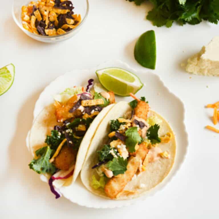 2 tacos made with fish sticks on a white plate topped with cilantro and cotija cheese.