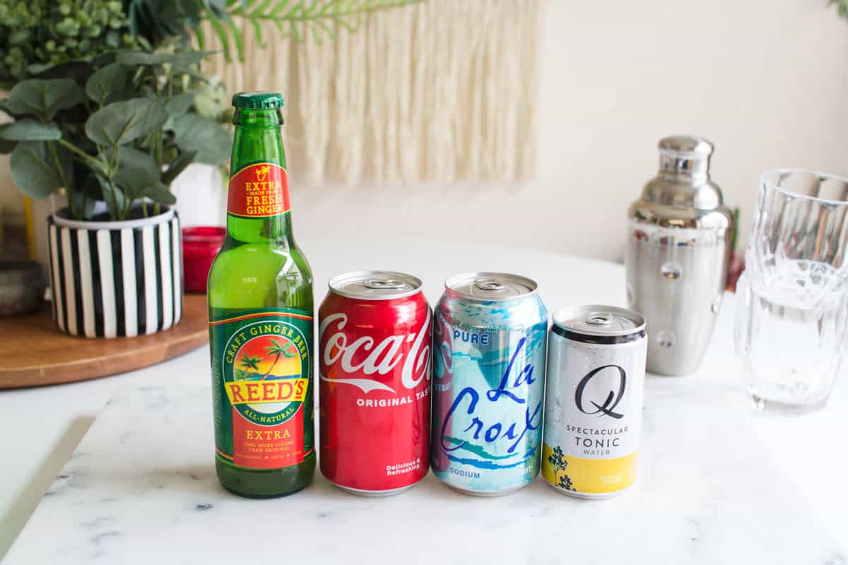 A variety of soda and tonic in bottles and cans on a table next to a cocktail shaker. 