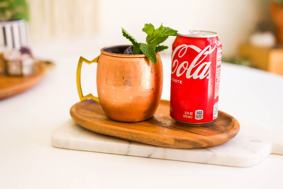 Close up of a copper Moscow Mule mug next to a can of Coke.