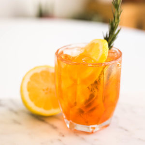 An orange cocktail in a cocktail glass with rosemary sprig.