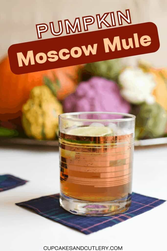 Text: Pumpkin Moscow Mule with a cocktail glass holding a cocktail on a table with a pumpkin centerpiece in the background.