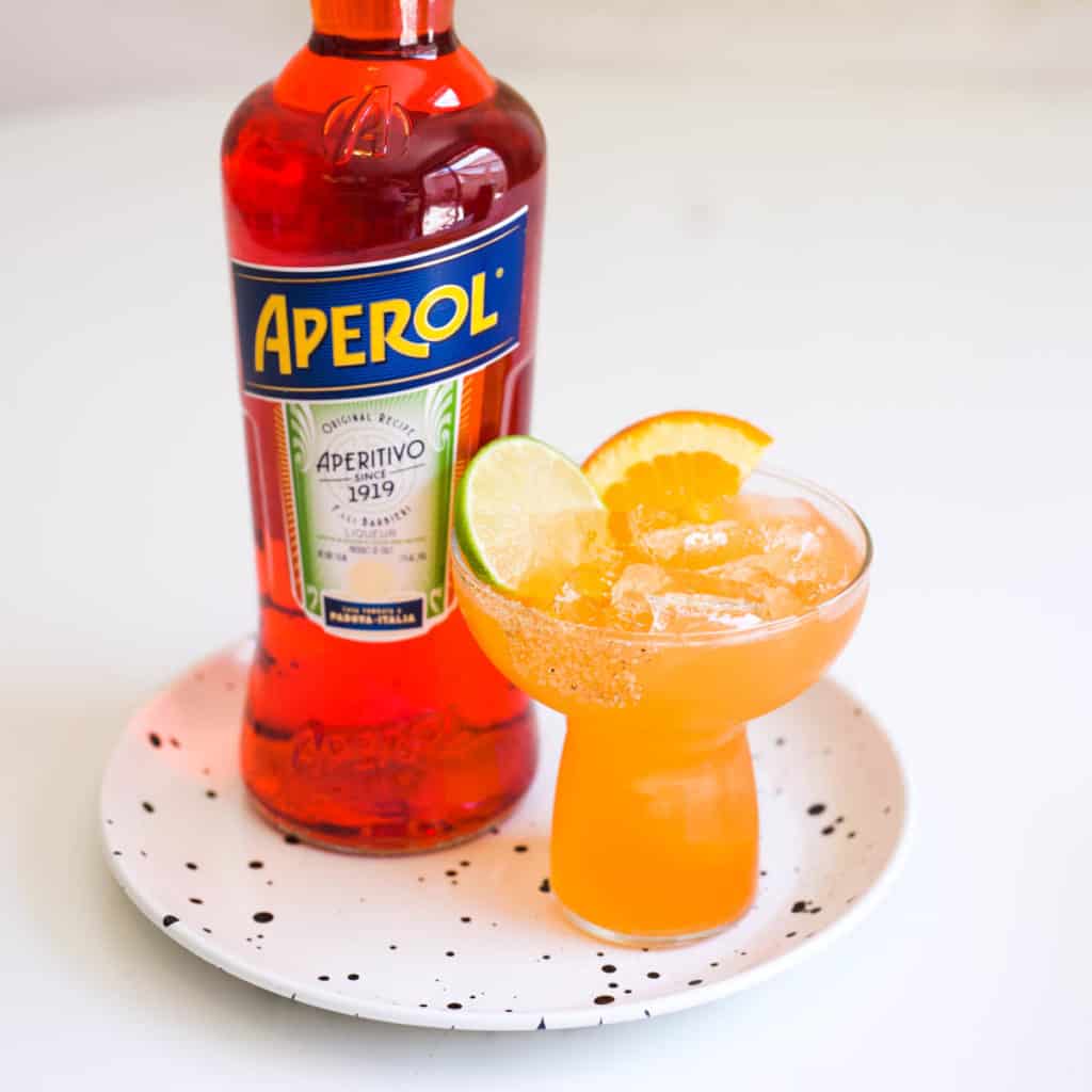 An Aperol Margarita in a stemless margarita glass on a small tray next to a bottle of Aperol.