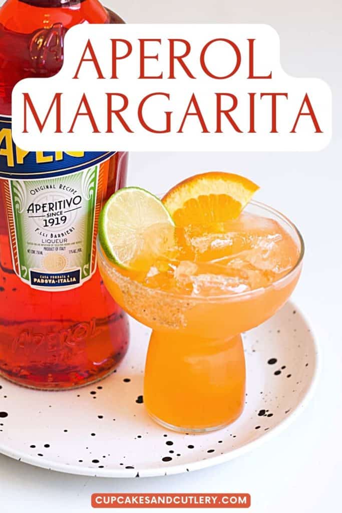 A stemless margarita glass on a table next to a bottle of Aperol with text that says "Aperol Margarita" on it.