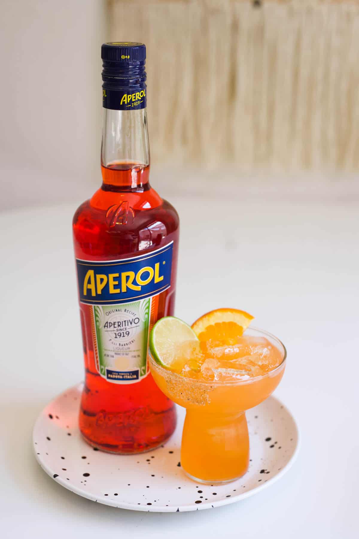 A bottle of Aperol on a small tray next to a cocktail glass with an Aperol Margarita.