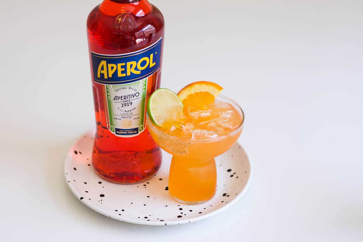 An orange margarita in a glass next to a bottle of Aperol. 