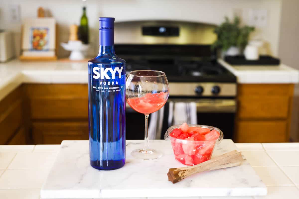 A bottle of Skyy vodka on a counter next to a wine glass with muddled watermelon in it.