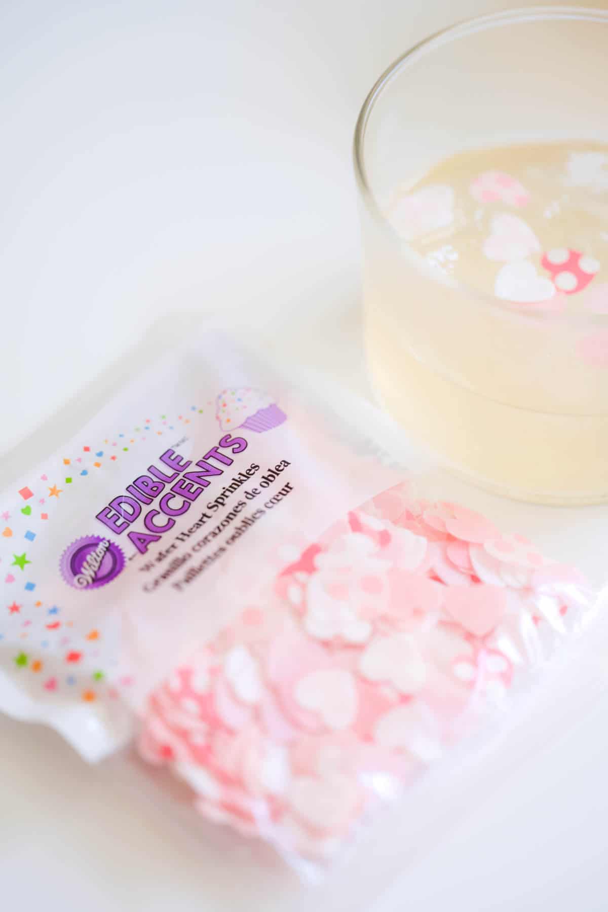 A package of edible confetti on a table next to a glass with a drink topped with the heart confetti.