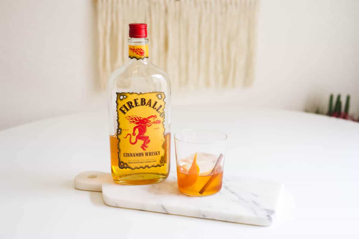 A bottle of Cinnamon Whiskey on a tray next to a cocktail glass with a Fireball Old Fashioned.