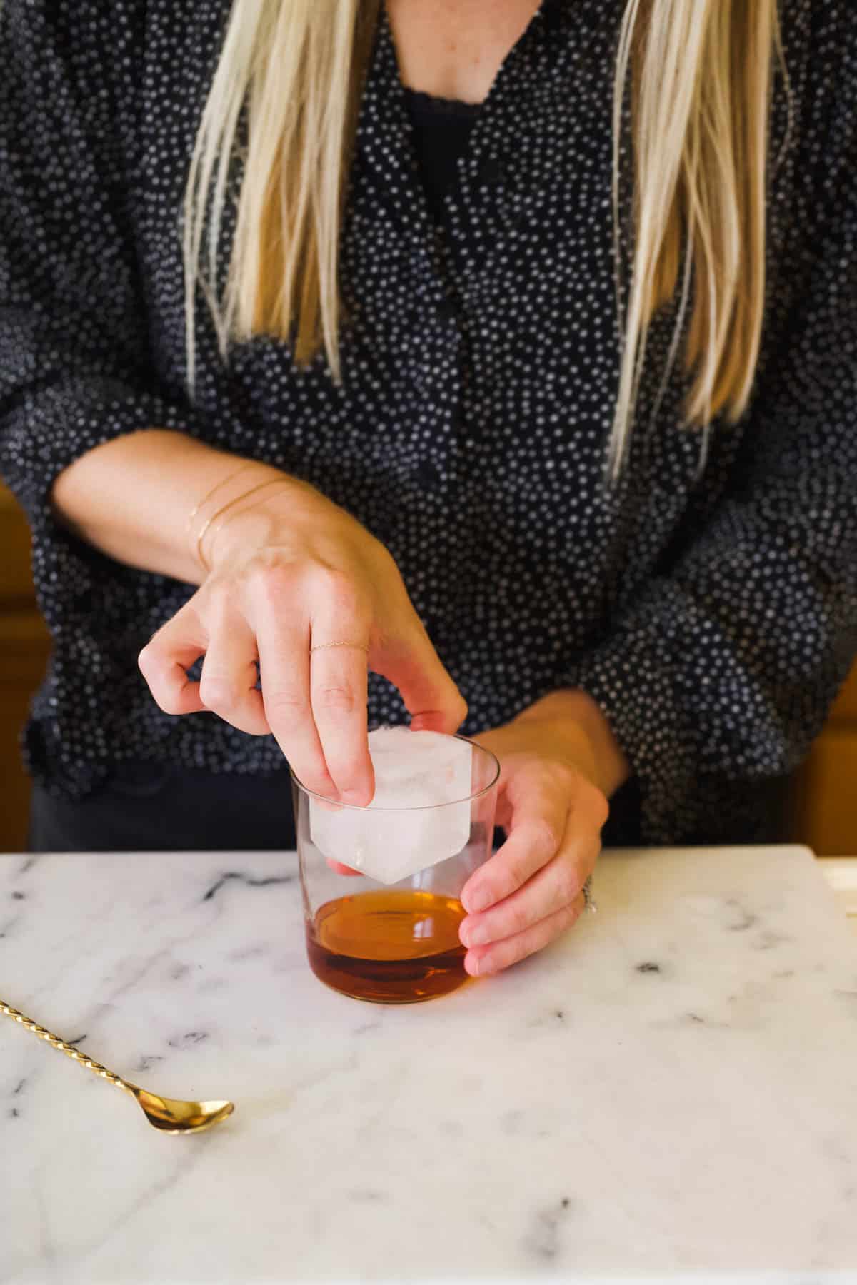 Woman adding a large ice cube to a cocktail glass for a cinnamon whiskey old fashioned.