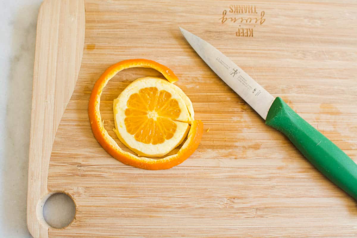 A wooden cutting board with a pairing knife and a slice of orange with the peel cut off of it.