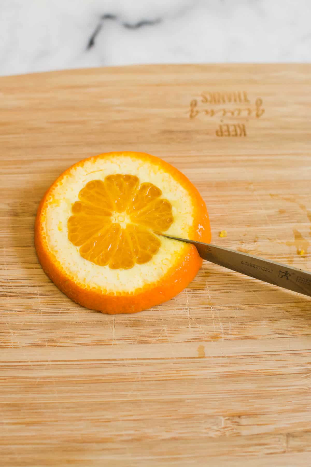An orange slice on a wooden cutting board with a pairing knife slicing through the peel.
