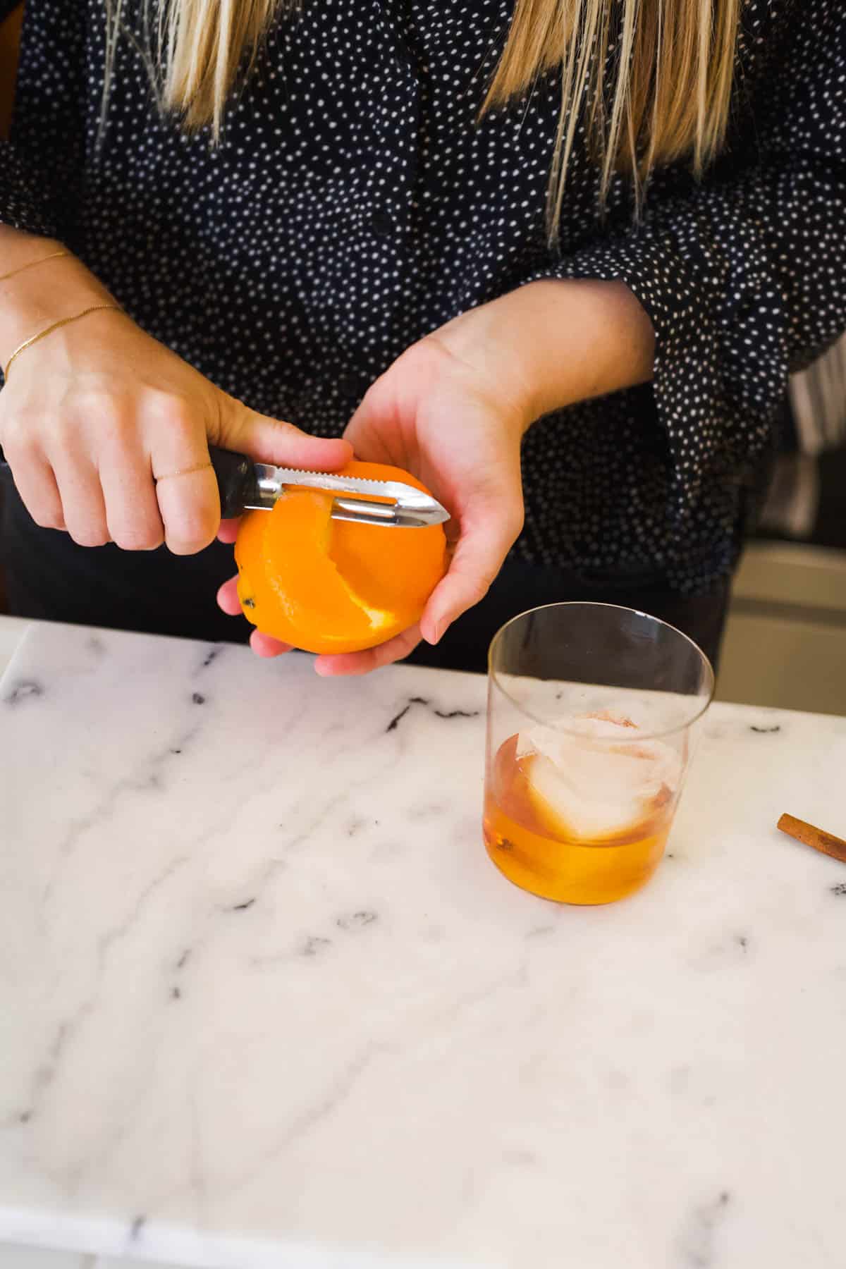 Woman peeling an orange with a vegetable peeler for an Old Fashioned garnish.