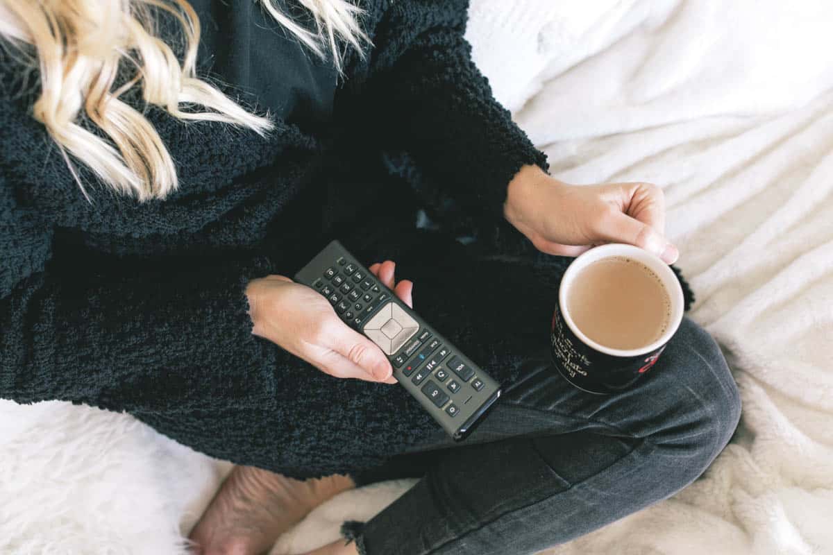 A curled up woman on the couch with coffee and a remote control for the TV. 