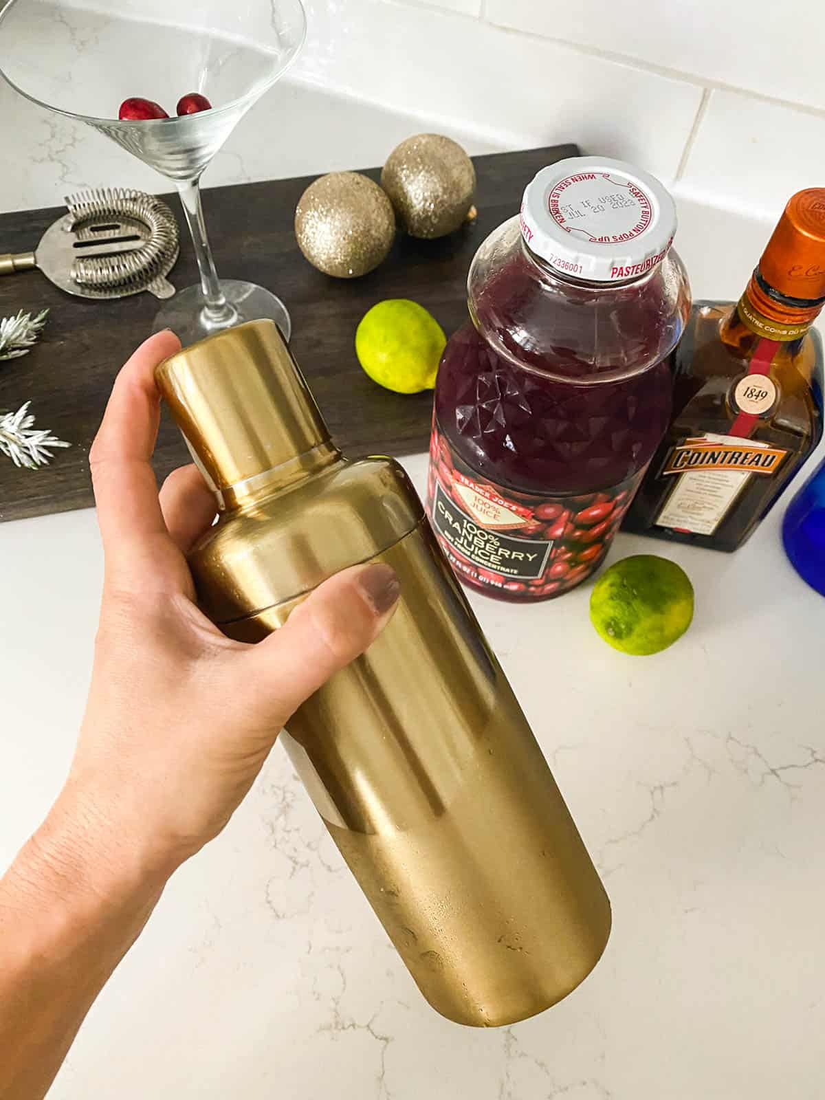 Woman shaking a gold cocktail shaker with cocktail ingredients in the background.