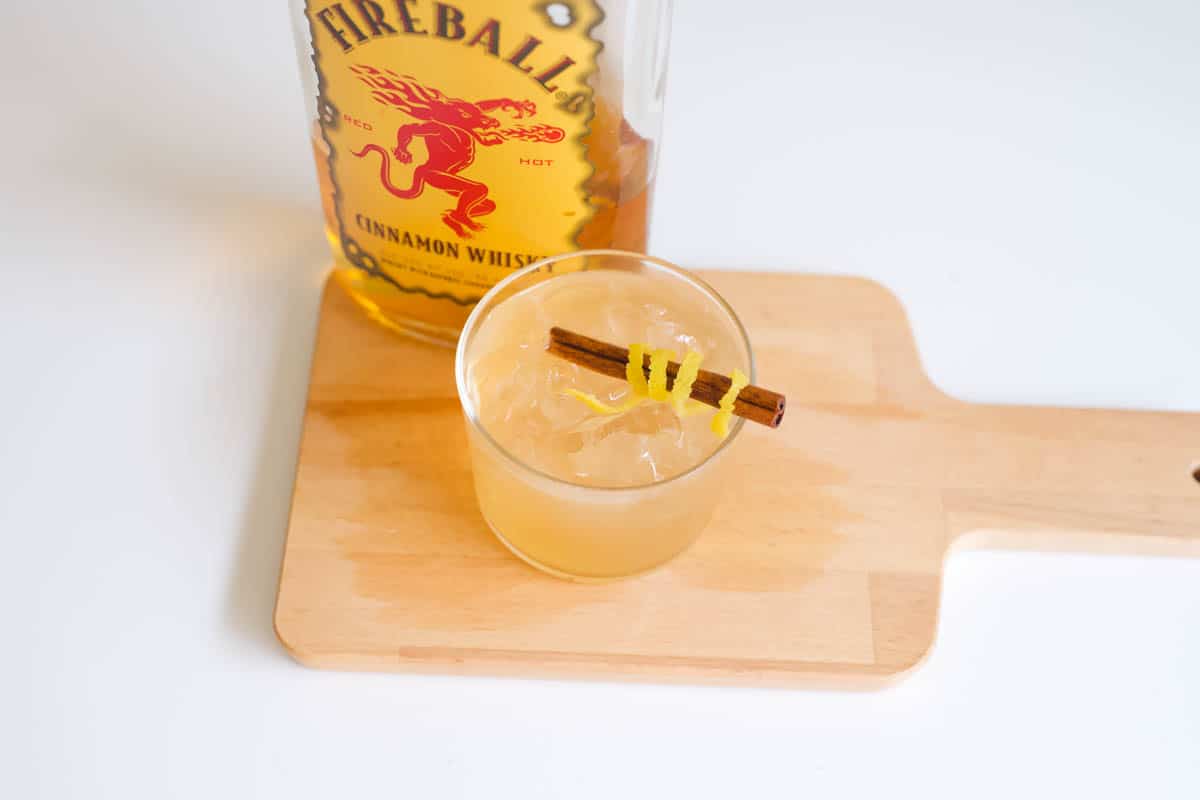A close up of a cinnamon whisky sour on a wooden board with Fireball near it. 