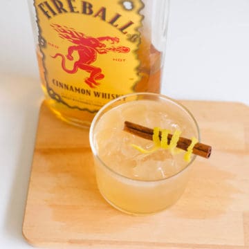 Close up of a Fireball Sour cocktail in a glass with a lemon peel wrapped cinnamon stick and a bottle of Fireball behind it.