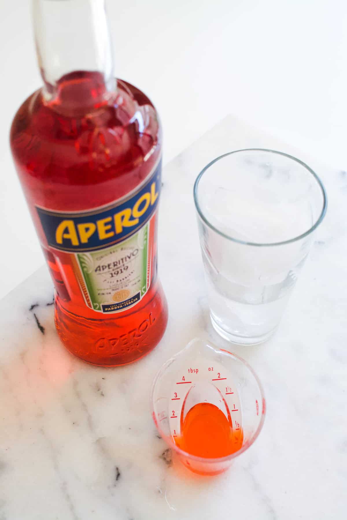 A bottle of Aperol on a table next to a glass of ice and a jigger measuring Aperol.