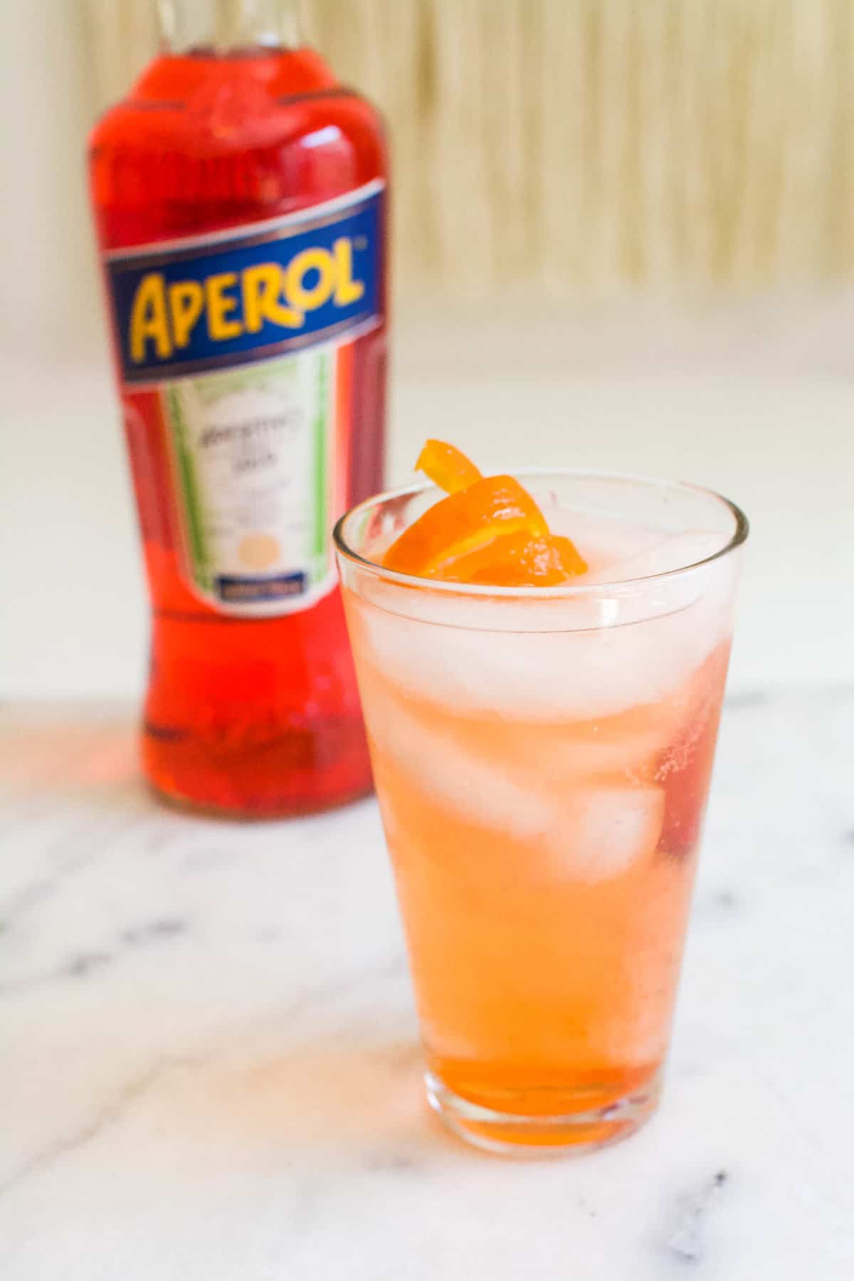 An Aperol Gin and Tonic over ice in a tall glass on a table with a garnish next to a bottle of Aperol.