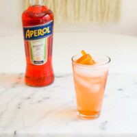 Aperol cocktail in a tall glass with ice on a table next to a bottle of Aperol.