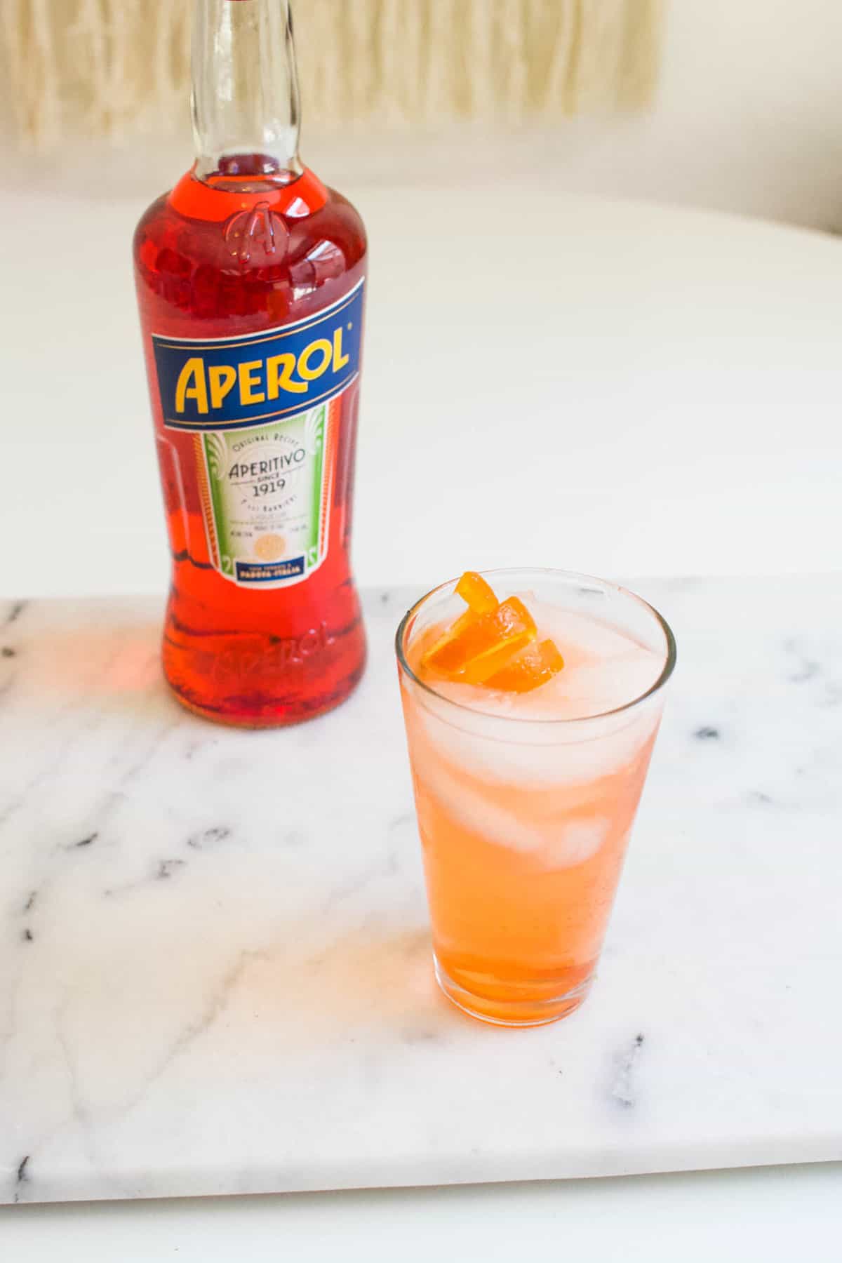 Orange peel spiral garnished cocktail on a table next to a bottle of Aperol.