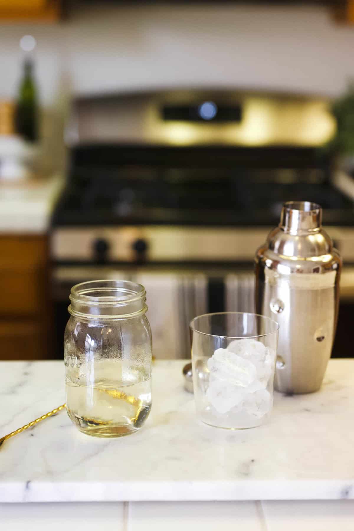 A jar of simple syrup on a counter next to a cocktail shaker and a short glass with ice.