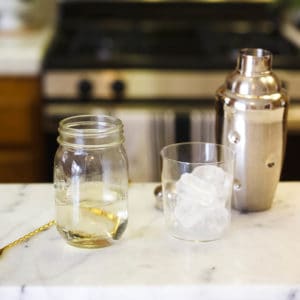 Mason jar holding simple syrup made in the microwave on a counter next to a glass of ice and a cocktail shaker.